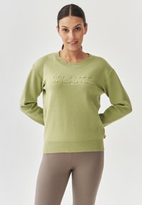 Suéter de color verde claro con cuello y puños acanalados, que presenta la palabra "BALANCE" en relieve en el frente. Textura de tela suave.