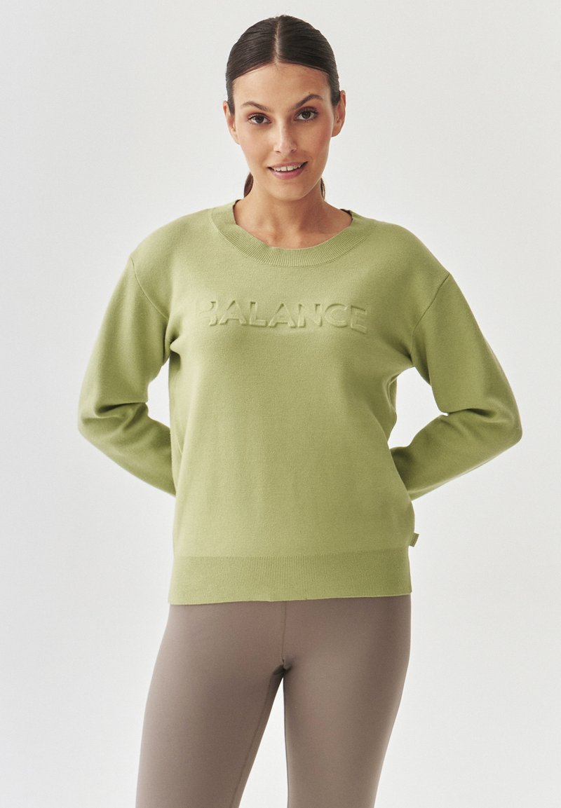 Suéter de color verde claro con cuello y puños acanalados, que presenta la palabra "BALANCE" en relieve en el frente. Textura de tela suave.