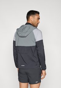 Ανδρικό μπουφάν με χρωματικές αντιθέσεις, γκρι κουκούλα, σκούρο μπλε σώμα και αντανακλαστικές λεπτομέρειες· ελαφρύ ύφασμα και casual εφαρμογή.