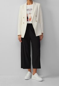 Witte dubbelzijdige blazer, graphic tee met "LIFE IS", zwarte gepliste culottes en witte sneakers. Glad materiaal, afgetailleerde pasvorm.