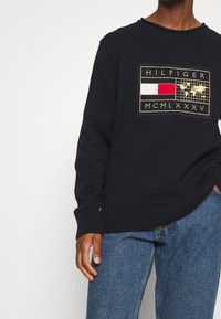 Maglione nero lavorato a maglia con una toppa del logo Tommy Hilfiger nei colori rosso, bianco e oro, indossato con jeans blu.