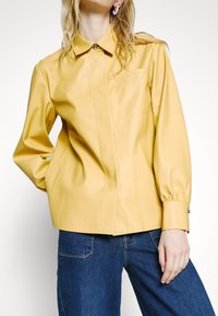 Femme portant une chemise jaune à manches longues boutonnée avec un col et un jean bleu, debout avec une main dans la poche et l'autre détendue.