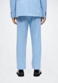 Άνδρας που φοράει ανοιχτό μπλε παντελόνι και σακάκι, στέκεται με μαύρα επίσημα παπούτσια, εμφανίζεται από πίσω σε λευκό φόντο.