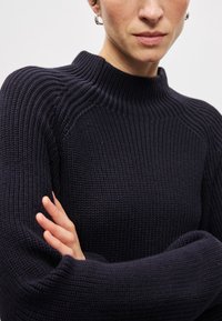 Ναυτικό ριμπ ελβετικό πουλόβερ με ψηλό λαιμό και χαλαρή εφαρμογή. Διαθέτει υφασμάτινη υφή και μακριά μανίκια με φαρδιές μανσέτες.