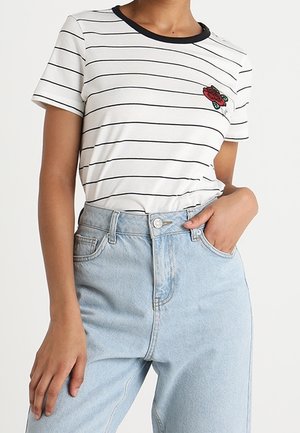 Donna che indossa una t-shirt bianca a righe con colletto nero e ricamo di rosa, infilata in jeans azzurri a vita alta.