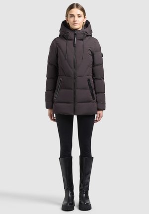 Jeune femme portant une veste d’hiver noire matelassée avec capuche, des leggings noirs et des bottes noires montant jusqu’aux genoux, debout devant un fond gris uni.