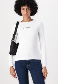 Biely top s dlhým rukávom a rebrovanou textúrou, s výšivkou "guessjeans"; skombinovaný s tmavomodrými džínsami a čiernou kabelkou cez plece.