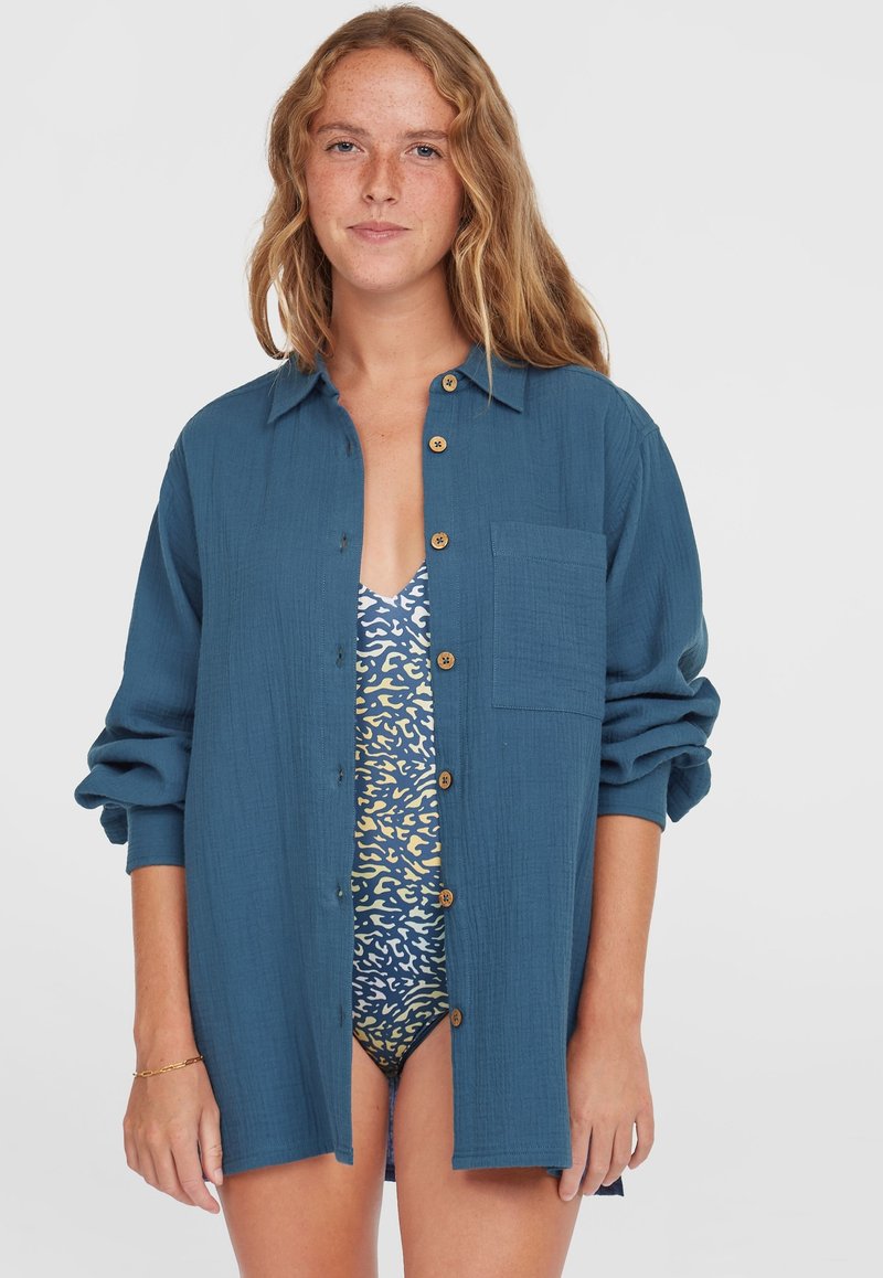 Blauwe katoenen button-up shirt met een zak, opgerolde mouwen en houten knopen, gedragen over een gestreept zwemkostuum in tinten van blauw en geel.