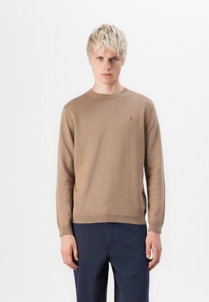 Brown knit sweater with a crew neck and ribbed cuffs. Features a small, subtle logo on the chest. Paired with dark navy trousers.