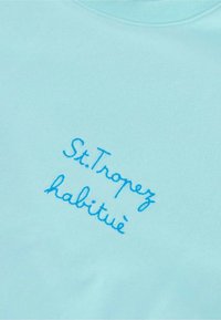 Pullover azzurro chiaro con testo ricamato; la scritta recita "St. Tropez habitué." Tessuto liscio, design con scollatura a girocollo.