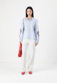 Camisa azul claro com designs florais pretos bordados nas mangas, combinada com calças brancas, uma bolsa vermelha e saltos vermelhos com detalhes metálicos.