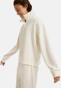 Pull crème avec un col haut, des manches longues et une coupe décontractée. Matière douce et détail de logo subtil sur la poitrine.