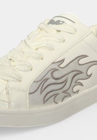 Close-up of a white sneaker with thick white laces and gray flame patterns on the side panel above a textured rubber sole.