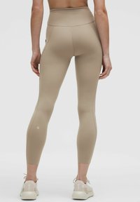 Beige, magas derekú leggings sima, rugalmas anyagból, oldalsó zsebekkel és logóval a bokánál. A fazon testhez simuló.