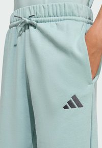 Svetlozelené tepláky s elastickým pásom, nastaviteľnou šnúrkou, bočnou vreckom a čiernym trojuholníkom loga Adidas. Mäkká textúra látky.