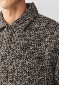 Chaqueta texturada en gris y marrón con cuello de solapa, que cuenta con cierres de botón y un patrón tejido de diferentes colores de hilo.