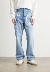 Light blue denim jeans with a relaxed fit, featuring varied blue embroidered designs and patches, including a smiley face and text labels.