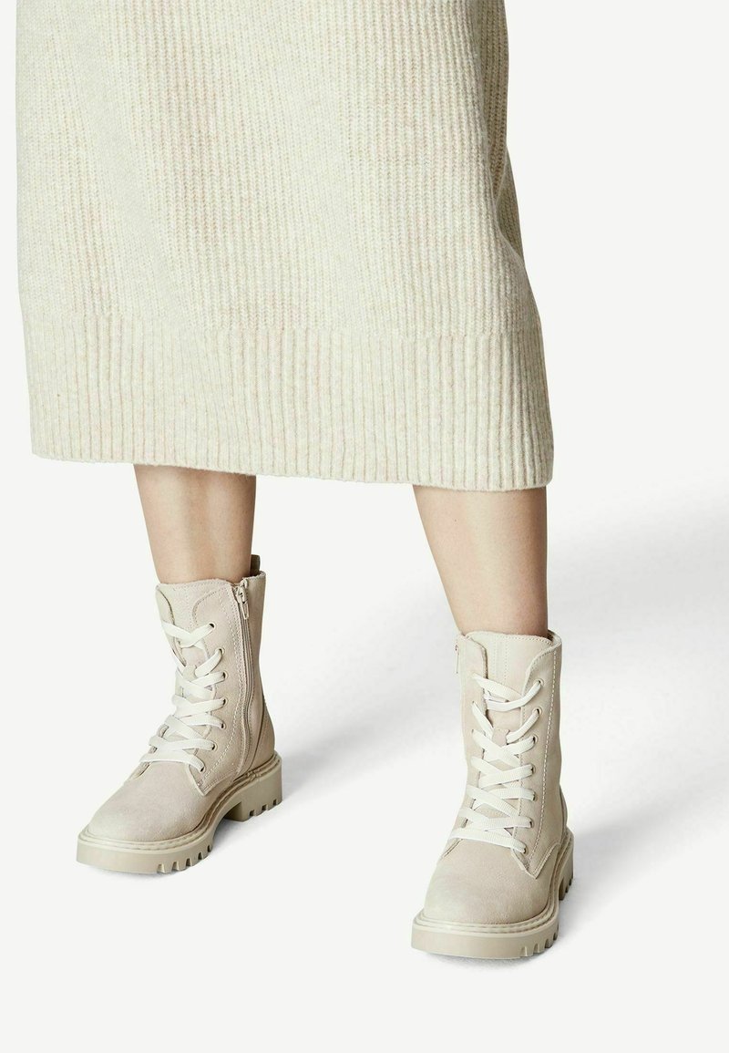 Femme portant une jupe midi en tricot crème et des bottes de combat à lacets beige clair avec des fermetures éclair latérales sur fond blanc.
