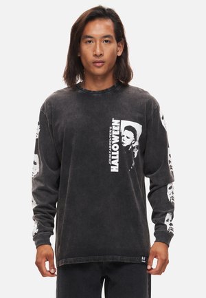 Chemise à manches longues, noir délavé, présentant des graphiques blancs, y compris un visage et du texte, avec des motifs de crâne sur les manches.