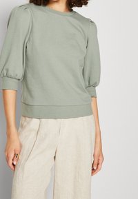 Grøn sweatshirt med puffede ærmer og rund halsudskæring. Blødt stof, tætsiddende i taljen, parret med lyse beige løse bukser.