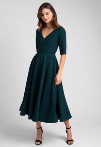 Sukienka o długości do kolan, w głębokim zielonym kolorze, z dopasowanym gorsetem, dekoltem w literę V i rękawami 3/4; rozkloszowany dół o gładkiej fakturze. Czarne sandały na kostkę.