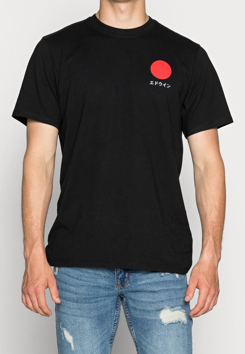 Homme portant un t-shirt noir à manches courtes avec un cercle rouge et un texte japonais blanc sur la poitrine, assorti à un jean bleu déchiré.