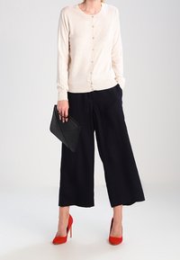 Beige långärmad kofta med knappar, i kombination med svarta vida byxor och röda högklackade skor. Håller en svart clutch.