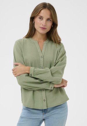 Kvinde med lysbrunt hår iført grøn knapbluse og lyseblå jeans, stående med armene krydset mod hvid baggrund.