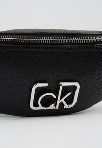 Pochette en cuir noir texturé avec fermeture éclair argentée et logo métallique « ck » sur le devant.