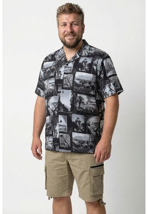 Lächelnder Mann mit Bart, der ein schwarz-weiß gemustertes kurzärmeliges Hemd und beigefarbene Cargo-Shorts trägt, steht vor einem schlichten weißen Hintergrund.