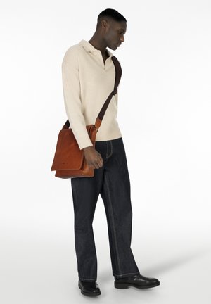 Pullover beige a maglia, jeans in denim scuro e borsa a tracolla in pelle marrone con cinturino regolabile; il modello è in posizione laterale contro uno sfondo bianco.