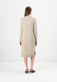 Abito in maglia beige in lana con maniche lunghe, orlo a coste e spacchi laterali. Presenta un colletto classico e un design al ginocchio. Indossato con tacchi neri.