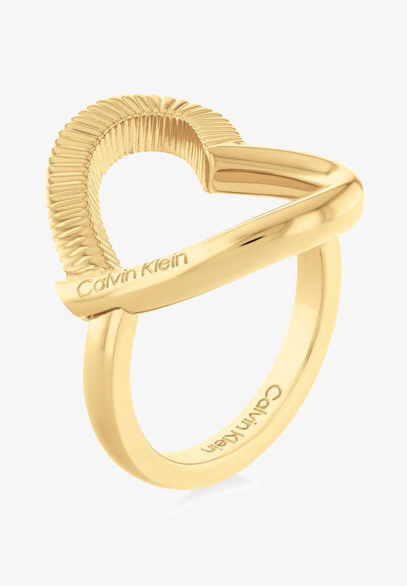 Anillo de color dorado con un diseño de corazón texturizado. Presenta bordes lisos y acanalados, con el nombre de la marca "Calvin Klein" grabado en la banda.