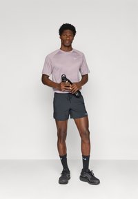 Ανοιχτό μωβ αθλητικό μπλουζάκι κοντομάνικο με υφασμάτινη υφή, συνδυασμένο με μαύρες βερμούδες και μαύρα αθλητικά παπούτσια. Κρατάει ένα μαύρο μπουκάλι νερού.