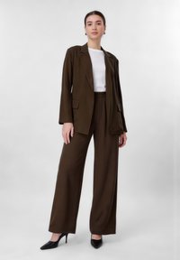 Femme debout portant un costume marron avec pantalon large, un haut blanc, des talons noirs et des boucles d'oreilles créoles dorées sur un fond gris uni.