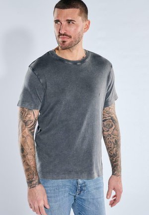 Man with short hair and full arm tattoos wearing a faded gray t-shirt and light blue jeans, looking to the left.