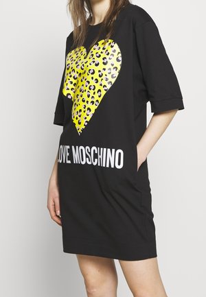 Nainen pukeutuneena mustaan mekkoon, jossa on suuri keltainen leopardikuvioinen sydän ja valkoinen "LOVE MOSCHINO" -teksti, kädet taskuissa, neutraali tausta.