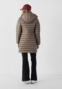 Gevoerde bruine jas met capuchon, voorzien van een horizontaal gestikt ontwerp, gestyled met zwarte flared broeken en een bordeauxrode pet, staand op een neutrale achtergrond.