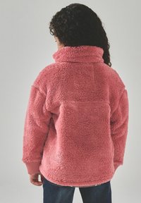 Person med krøllet hår iført en løs, lyserød, blød jakke set bagfra mod en ensfarvet baggrund.