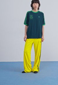 Green and navy Adidas t-shirt with green accents, paired with loose-fit yellow track pants featuring black side stripes. Soft fabric, casual design.