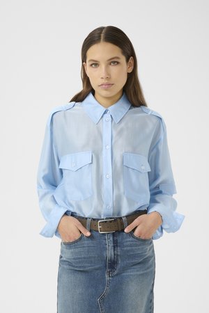 Woman with brown hair wearing a light blue button-up shirt with chest pockets and a denim skirt with a brown belt, hands in pockets.