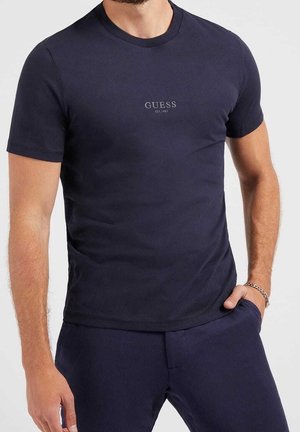 Homme portant un t-shirt ajusté bleu marine foncé à manches courtes avec le texte "GUESS EST. 1981" centré sur la poitrine et un pantalon bleu marine assorti.