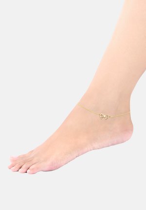 Goldene Fußkette mit einer zarten Kette und einem schleifenförmigen Anhänger. Glatte Metalloberfläche mit glänzendem Finish. Am baren Fuß getragen.