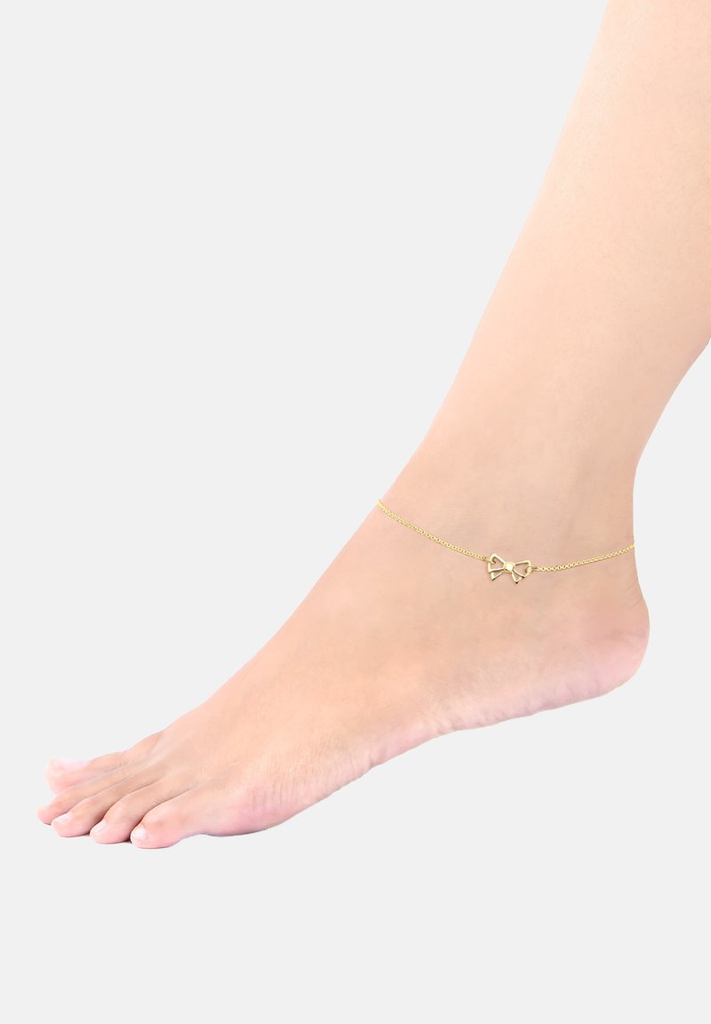 Guldanklet med en delikat kæde og en sløjfeformet charm. Glat metalsurface med en blank finish. Båret på en bar fod.