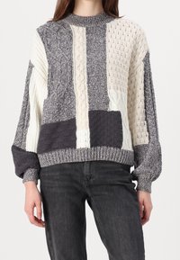 Suéter de punto con un diseño de patchwork en gris, crema y gris oscuro. Los patrones texturizados incluyen trenzas y punto acanalado, con un cuello alto.