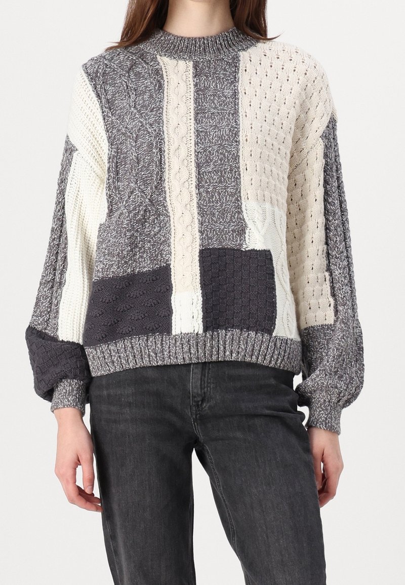 Suéter de punto con un diseño de patchwork en gris, crema y gris oscuro. Los patrones texturizados incluyen trenzas y punto acanalado, con un cuello alto.