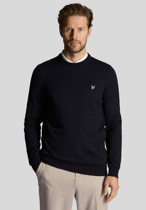 Mand med lyst brunt hår og skæg iført en sort rundhalset sweater med et lille grønt logo og beige bukser, der står mod en grå baggrund.