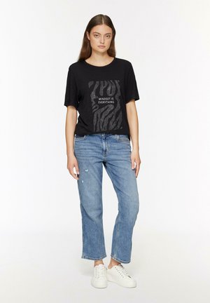 T-shirt noir à manches courtes avec un graphisme texturé en argent portant l'inscription "L'ÉTAT D'ESPRIT EST TOUT." Accompagné d'un jean déchiré bleu clair et de baskets blanches.