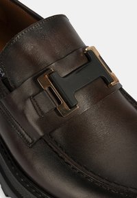 Braune Lederloafers mit glänzender Oberfläche, ausgestattet mit einer gold-schwarzen dekorativen Schnalle über dem Rist und sichtbaren Nähakzenten.