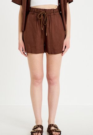 Brune linnedragtshorts med snøre brugt med hvid cropped top og mørkebrune sandaler med dobbelt spænde på model stående mod hvid baggrund.